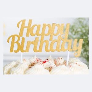 Gold metallic "Happy Birthday" cake topper inserted into white frosted cake with decorative swirls, creating an elegant birthday party centerpiece against a soft background