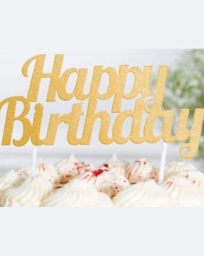 Gold metallic "Happy Birthday" cake topper inserted into white frosted cake with decorative swirls, creating an elegant birthday party centerpiece against a soft background