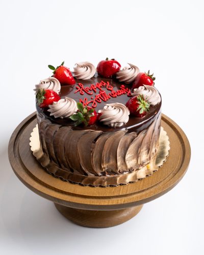 7 Layer Birthday Cake from Mia's Bakery featuring rich chocolate layers with glossy chocolate drip frosting on the sides. The birthday cake is topped with alternating swirls of chocolate and vanilla buttercream rosettes, fresh glazed strawberries, and a red 'Happy Birthday!' message written in cursive on the chocolate surface. Displayed on a golden pedestal cake stand against a white background.