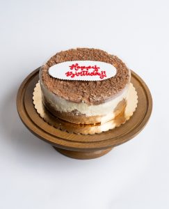 Apple Cheesecake Birthday Cake from Mia's Bakery featuring a round layered cake with creamy white cheesecake filling, golden cake base, and brown cinnamon apple crumble topping. Decorated with a white fondant plaque reading 'Happy Birthday!' in red cursive lettering. The birthday cake is presented on a wooden pedestal cake stand against a white background.