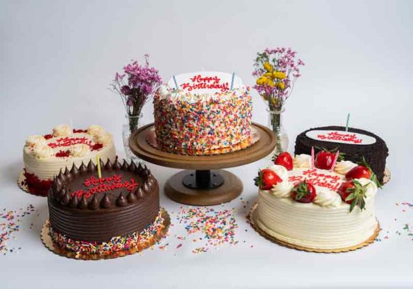 Three birthday cakes from Mia's Bakery displayed on stands: a rich chocolate birthday cake with chocolate frosting and red Happy Birthday topper on the left, a colorful confetti sprinkle birthday cake with red candles and Happy Birthday topper in the center on a raised pedestal, and an elegant white frosted birthday cake with fresh strawberries and Happy Birthday decoration on the right. Purple and pink flower arrangements in vases complement the festive cake display.