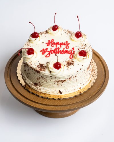 Black Forest Birthday Cake from Mia's Bakery featuring a round chocolate layer cake with white whipped cream frosting and horizontal ridged texture. The birthday cake is decorated with piped whipped cream rosettes topped with maraschino cherries with stems around the top edge. Chocolate shavings are dusted over the entire surface. 'Happy Birthday!' is written in red cursive frosting in the center. Presented on a wooden pedestal cake stand against a white background.