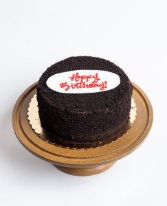 Blackout Birthday Cake from Mia's Bakery featuring a round chocolate layer cake completely covered in dark chocolate crumbs. The birthday cake has chocolate cake crumbs pressed onto all surfaces creating an all-black appearance. 'Happy Birthday!' is written in red cursive on a white fondant plaque placed on top. The cake sits on a golden cake board. Displayed on a wooden pedestal cake stand against a white background.