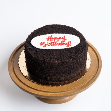 Blackout Birthday Cake from Mia's Bakery featuring a round chocolate layer cake completely covered in dark chocolate crumbs. The birthday cake has chocolate cake crumbs pressed onto all surfaces creating an all-black appearance. 'Happy Birthday!' is written in red cursive on a white fondant plaque placed on top. The cake sits on a golden cake board. Displayed on a wooden pedestal cake stand against a white background.