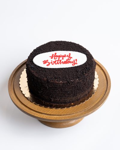 Blackout Birthday Cake from Mia's Bakery featuring a round chocolate layer cake completely covered in dark chocolate crumbs. The birthday cake has chocolate cake crumbs pressed onto all surfaces creating an all-black appearance. 'Happy Birthday!' is written in red cursive on a white fondant plaque placed on top. The cake sits on a golden cake board. Displayed on a wooden pedestal cake stand against a white background.