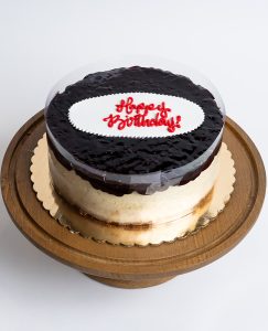 Blueberry Cheesecake Birthday Cake from Mia's Bakery featuring a round cheesecake with creamy white sides and glossy dark blueberry glaze topping. The birthday cake has a deep purple-blue blueberry glaze with visible berry texture covering the top surface. 'Happy Birthday!' is written in red cursive on a white fondant plaque centered on the blueberry topping. The cake has a clear acetate collar around the edge. Displayed on a wooden pedestal cake stand against a white background.