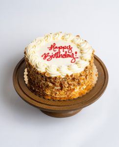 Burnt Almond Birthday Cake from Mia's Bakery featuring a round layer cake with white buttercream frosting and toasted sliced almonds completely covering the sides. The birthday cake is decorated with piped white buttercream rosettes around the top edge. 'Happy Birthday!' is written in red cursive frosting in the center. The cake has a golden caramel drizzle visible at the base. Presented on a wooden pedestal cake stand against a white background.