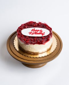 Cherry Cheesecake Birthday Cake from Mia's Bakery featuring a round cheesecake with creamy white sides and glossy red cherry topping. The birthday cake has a ruby-red cherry glaze with visible cherry pieces covering the top surface. 'Happy Birthday!' is written in red cursive on a white fondant plaque centered on the cherry topping. The cake shows a golden graham cracker crust base. Presented on a wooden pedestal cake stand against a white background.