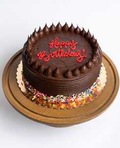 Chocolate Fudge Birthday Cake from Mia's Bakery featuring a round chocolate layer cake completely covered in rich chocolate fudge frosting with horizontal ridged texture on the sides. The birthday cake is decorated with piped chocolate buttercream kisses arranged in a border around the top edge. 'Happy Birthday!' is written in red cursive directly on the chocolate surface. Rainbow sprinkles decorate the bottom edge. Presented on a wooden pedestal cake stand against a white background.