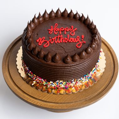 Chocolate Fudge Birthday Cake from Mia's Bakery featuring a round chocolate layer cake completely covered in rich chocolate fudge frosting with horizontal ridged texture on the sides. The birthday cake is decorated with piped chocolate buttercream kisses arranged in a border around the top edge. 'Happy Birthday!' is written in red cursive directly on the chocolate surface. Rainbow sprinkles decorate the bottom edge. Presented on a wooden pedestal cake stand against a white background.