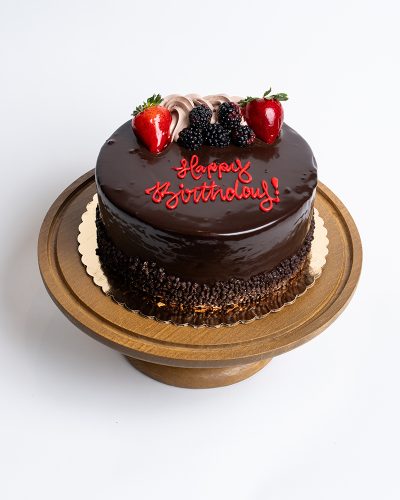 Chocolate Layer Birthday Cake from Mia's Bakery featuring a round cake with glossy dark chocolate ganache coating the entire surface. The birthday cake is decorated with fresh strawberries, blackberries, and chocolate buttercream rosettes on top. 'Happy Birthday!' is written in red cursive frosting directly on the chocolate surface. The cake has chocolate pearls or sprinkles around the bottom edge. Displayed on a wooden pedestal cake stand against a white background.