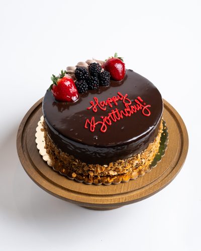 Chocolate Mousse Birthday Cake from Mia's Bakery featuring a round cake with glossy dark chocolate mirror glaze covering the entire top and sides. The birthday cake is decorated with fresh strawberries, blackberries, and chocolate shavings on top, with crushed almonds pressed around the bottom edge. 'Happy Birthday!' is written in red cursive frosting directly on the chocolate surface. Presented on a wooden pedestal cake stand against a white background.