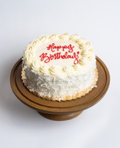 Coconut Birthday Cake from Mia's Bakery featuring a round single-layer cake completely covered in white buttercream with shredded coconut flakes pressed onto the sides. The birthday cake is decorated with piped vanilla buttercream rosettes around the top edge and displays 'Happy Birthday!' written in red cursive frosting in the center. Presented on a wooden pedestal cake stand against a white background.
