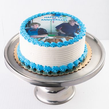 Custom photo edible image anniversary cake from Mia's Bakery featuring a round white layer cake with horizontal ridged buttercream texture on the sides. The anniversary cake displays a circular edible image print showing a couple's photo with 'HAPPY 50TH ANNIVERSARY' text at the top. Blue buttercream rosettes border the edible image and decorate the bottom edge. Presented on a metallic cake stand against a white background.
