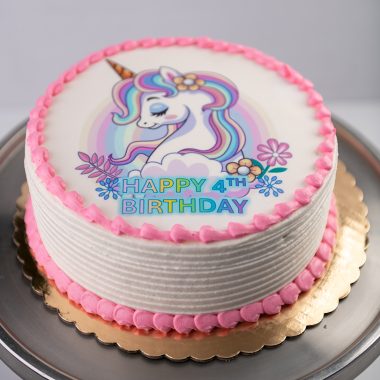 Edible Image Birthday Cake from Mia's Bakery featuring a round white layer cake with horizontal ridged buttercream texture on the sides. The birthday cake is decorated with a circular edible image print of a magical unicorn with rainbow mane and purple flowers on top. Pink buttercream pearls border the edible image and the bottom edge of the cake. 'HAPPY 4TH BIRTHDAY' is printed in purple text on the edible image. Displayed on a gold cake board against a gray background.