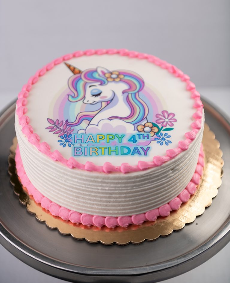 Edible Image Birthday Cake from Mia's Bakery featuring a round white layer cake with horizontal ridged buttercream texture on the sides. The birthday cake is decorated with a circular edible image print of a magical unicorn with rainbow mane and purple flowers on top. Pink buttercream pearls border the edible image and the bottom edge of the cake. 'HAPPY 4TH BIRTHDAY' is printed in purple text on the edible image. Displayed on a gold cake board against a gray background.