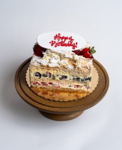 Fruit Napoleon Birthday Cake from Mia's Bakery featuring multiple layers of flaky puff pastry with visible layers containing fresh blueberries, strawberries, and pastry cream filling. The birthday cake is topped with powdered sugar, whipped cream dollops, and fresh strawberries. 'Happy Birthday!' is written in red cursive on a white fondant plaque placed on top. The Napoleon shows golden caramelized base. Presented on a wooden pedestal cake stand against a white background.