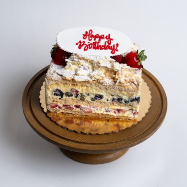 Fruit Napoleon Birthday Cake from Mia's Bakery featuring multiple layers of flaky puff pastry with visible layers containing fresh blueberries, strawberries, and pastry cream filling. The birthday cake is topped with powdered sugar, whipped cream dollops, and fresh strawberries. 'Happy Birthday!' is written in red cursive on a white fondant plaque placed on top. The Napoleon shows golden caramelized base. Presented on a wooden pedestal cake stand against a white background.