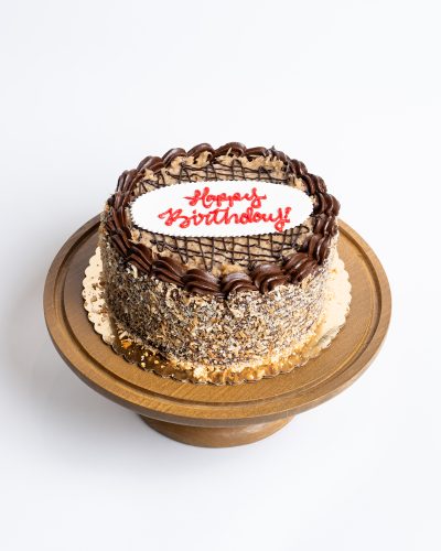 German Chocolate Birthday Cake from Mia's Bakery featuring a round layer cake with toasted coconut and chopped pecans completely covering the sides. The birthday cake is topped with signature caramel-colored coconut pecan frosting, chocolate drizzle pattern, and piped chocolate buttercream border around the edge. 'Happy Birthday!' is written in red cursive on a white fondant plaque in the center. Displayed on a wooden pedestal cake stand against a white background.