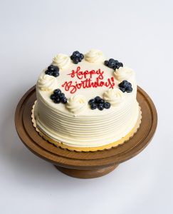 Lemon Blueberry Birthday Cake from Mia's Bakery featuring a round single-layer cake covered in smooth lemon-tinted white buttercream with horizontal ridged texture on the sides. The birthday cake is decorated with alternating fresh blueberry clusters and piped vanilla buttercream rosettes around the top edge. 'Happy Birthday!' is written in red cursive frosting in the center. Presented on a wooden pedestal cake stand against a white background.