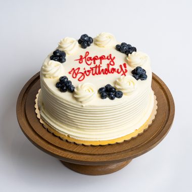 Lemon Blueberry Birthday Cake from Mia's Bakery featuring a round single-layer cake covered in smooth lemon-tinted white buttercream with horizontal ridged texture on the sides. The birthday cake is decorated with alternating fresh blueberry clusters and piped vanilla buttercream rosettes around the top edge. 'Happy Birthday!' is written in red cursive frosting in the center. Presented on a wooden pedestal cake stand against a white background.