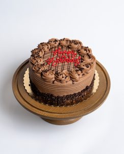 Nutella Birthday Cake from Mia's Bakery featuring a round chocolate layer cake completely covered in rich Nutella-style chocolate buttercream with horizontal ridged texture on the sides. The birthday cake is decorated with piped chocolate buttercream rosettes around the top edge and has chocolate crumbs pressed around the bottom edge. The top features chocolate drizzle lines and 'Happy Birthday!' written in red cursive frosting. Displayed on a wooden pedestal cake stand against a white background.