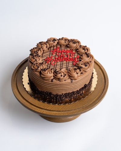 Nutella Birthday Cake from Mia's Bakery featuring a round chocolate layer cake completely covered in rich Nutella-style chocolate buttercream with horizontal ridged texture on the sides. The birthday cake is decorated with piped chocolate buttercream rosettes around the top edge and has chocolate crumbs pressed around the bottom edge. The top features chocolate drizzle lines and 'Happy Birthday!' written in red cursive frosting. Displayed on a wooden pedestal cake stand against a white background.