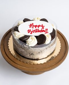 Oreo Cheesecake Birthday Cake from Mia's Bakery featuring a round cheesecake with white cream filling and visible Oreo cookie crumb layers on the sides. The birthday cake is decorated with whole and half Oreo cookies alternating with piped vanilla buttercream rosettes around the top edge. 'Happy Birthday!' is written in red cursive on a white fondant plaque in the center. The cake has a clear acetate collar around the sides. Displayed on a wooden pedestal cake stand against a white background.