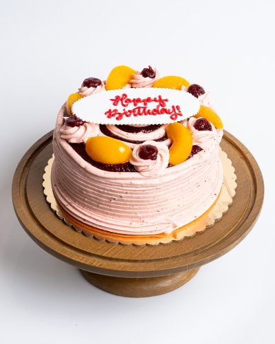 Peach Raspberry Birthday Cake from Mia's Bakery featuring a round single-layer cake covered in pink raspberry buttercream with horizontal ridged texture on the sides. The birthday cake is decorated with fresh peach slices, raspberry jam dollops, piped pink buttercream rosettes, and dark chocolate cake layers visible through decorative openings. 'Happy Birthday!' is written in red cursive on a white fondant plaque placed on top. Presented on a wooden pedestal cake stand against a white background.
