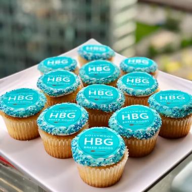 Dozen vanilla edible image cupcakes featuring HBG company logo printed on turquoise fondant toppers. These corporate photo cupcakes from Mia's Bakery display white "HBG" text on bright turquoise circular edible prints, topped with turquoise buttercream piped borders. Arranged on a white serving tray with city skyline backdrop, perfect for corporate events, business meetings, and company celebrations.