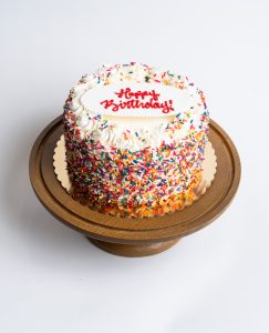Rainbow Birthday Cake from Mia's Bakery featuring a round single-layer cake covered in white vanilla buttercream with multicolored rainbow sprinkles completely coating the sides. The birthday cake is decorated with piped white buttercream rosettes around the top edge, each topped with rainbow sprinkles. 'Happy Birthday!' is written in red cursive frosting on a white fondant plaque centered on top. Displayed on a wooden pedestal cake stand against a white background.