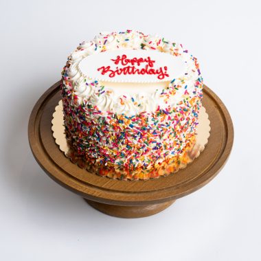 Rainbow Birthday Cake from Mia's Bakery featuring a round single-layer cake covered in white vanilla buttercream with multicolored rainbow sprinkles completely coating the sides. The birthday cake is decorated with piped white buttercream rosettes around the top edge, each topped with rainbow sprinkles. 'Happy Birthday!' is written in red cursive frosting on a white fondant plaque centered on top. Displayed on a wooden pedestal cake stand against a white background.