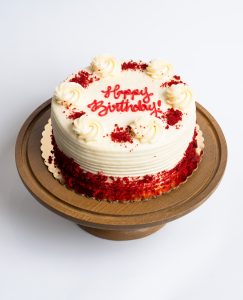Red Velvet Birthday Cake from Mia's Bakery featuring a round single-layer cake with white cream cheese frosting and horizontal ridged texture on the sides. The birthday cake has red velvet cake crumbs pressed around the bottom edge and is decorated with piped vanilla buttercream rosettes around the top edge, each sprinkled with red velvet cake crumbs. 'Happy Birthday!' is written in red cursive frosting in the center. Presented on a wooden pedestal cake stand against a white background.