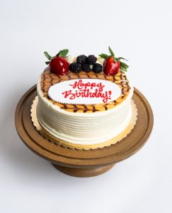 Salted Caramel Birthday Cake from Mia's Bakery featuring a round single-layer cake with white buttercream sides and horizontal ridged texture. The birthday cake is topped with golden salted caramel drizzle in a decorative pattern, fresh strawberries, blackberries, and piped white buttercream rosettes. 'Happy Birthday!' is written in red cursive on a white fondant plaque placed on the caramel-drizzled surface. Displayed on a wooden pedestal cake stand against a white background.