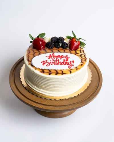 Salted Caramel Birthday Cake from Mia's Bakery featuring a round single-layer cake with white buttercream sides and horizontal ridged texture. The birthday cake is topped with golden salted caramel drizzle in a decorative pattern, fresh strawberries, blackberries, and piped white buttercream rosettes. 'Happy Birthday!' is written in red cursive on a white fondant plaque placed on the caramel-drizzled surface. Displayed on a wooden pedestal cake stand against a white background.