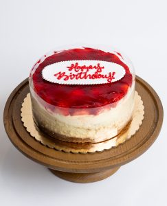 Strawberry Cheesecake Birthday Cake from Mia's Bakery featuring a round cheesecake with creamy white sides and glossy red strawberry glaze topping. The birthday cake displays 'Happy Birthday!' written in red cursive on a white fondant plaque centered on the strawberry glaze. The cake shows visible layers with a golden graham cracker crust base. Presented on a wooden pedestal cake stand against a white background.