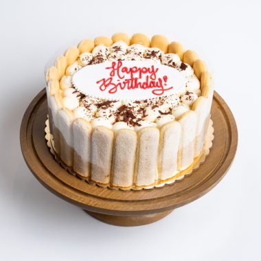 Tiramisu Birthday Cake from Mia's Bakery featuring a round cake decorated with ladyfinger cookies standing vertically around the sides. The birthday cake has a white cream top dusted with cocoa powder and chocolate shavings, with 'Happy Birthday!' written in red cursive frosting in the center on a white fondant plaque. Displayed on a wooden pedestal cake stand against a white background.