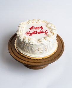 Vanilla Bean Birthday Cake from Mia's Bakery featuring a round single-layer cake covered in smooth white vanilla buttercream with horizontal ridged texture on the sides. The birthday cake is decorated with elegant piped buttercream rosettes and stars around the top edge and displays 'Happy Birthday!' written in red cursive frosting in the center. Presented on a wooden pedestal cake stand against a white background.