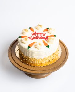 Carrot Birthday Cake from Mia's Bakery featuring a round single-layer cake covered in white cream cheese frosting with horizontal ridged texture on the sides. The birthday cake is decorated with piped buttercream rosettes around the top edge, each adorned with orange marzipan carrots and green leaves. 'Happy Birthday!' is written in red cursive in the center. Chopped walnuts are pressed around the bottom edge. Displayed on a wooden pedestal cake stand against a white background.