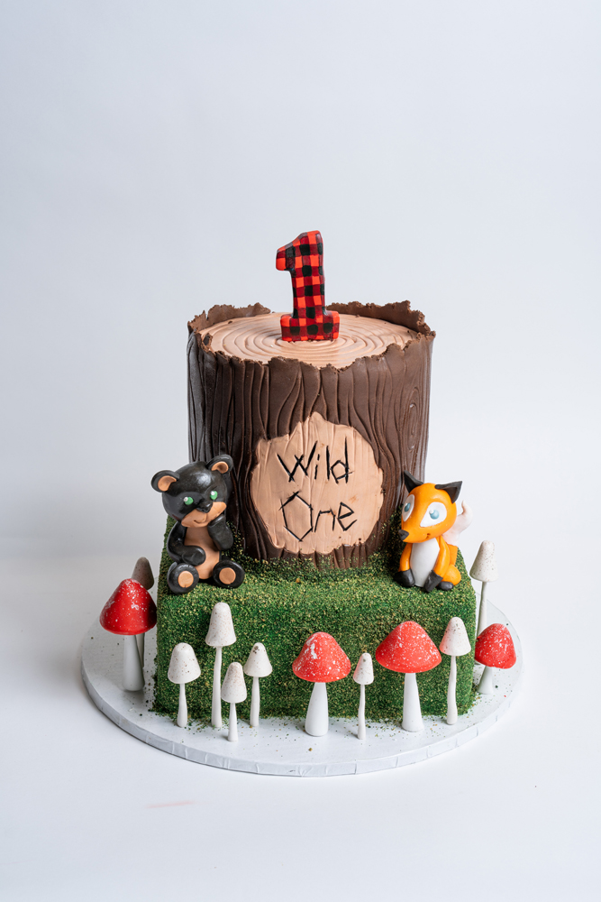 Custom lumberjack forest birthday cake from Mia's Bakery featuring two tiers designed as a tree stump with chocolate bark texture on top and green grass texture below. The cake displays 'Wild One' text on a wood slice plaque and features a red buffalo plaid number '1' on top. Decorated with fondant forest animals including a black bear and orange fox, plus red and white mushroom decorations around the bottom. Created for a first birthday celebration.