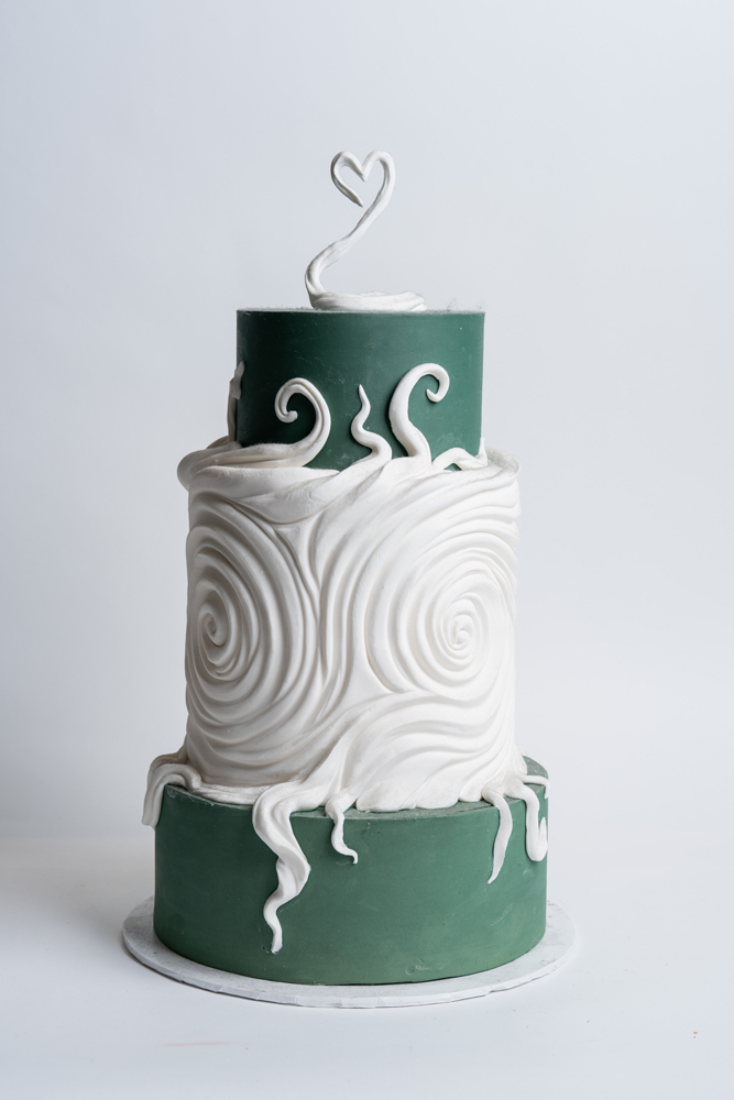 Custom swan-themed wedding cake from Mia's Bakery featuring three tiers alternating between dark sage green and white. The middle tier showcases dramatic white fondant swirls creating a wave pattern, while the green tiers feature elegant white swan designs. Topped with a silver swan heart cake topper. The sophisticated design includes flowing fondant waves connecting the tiers. Displayed on a white cake board against a white background.