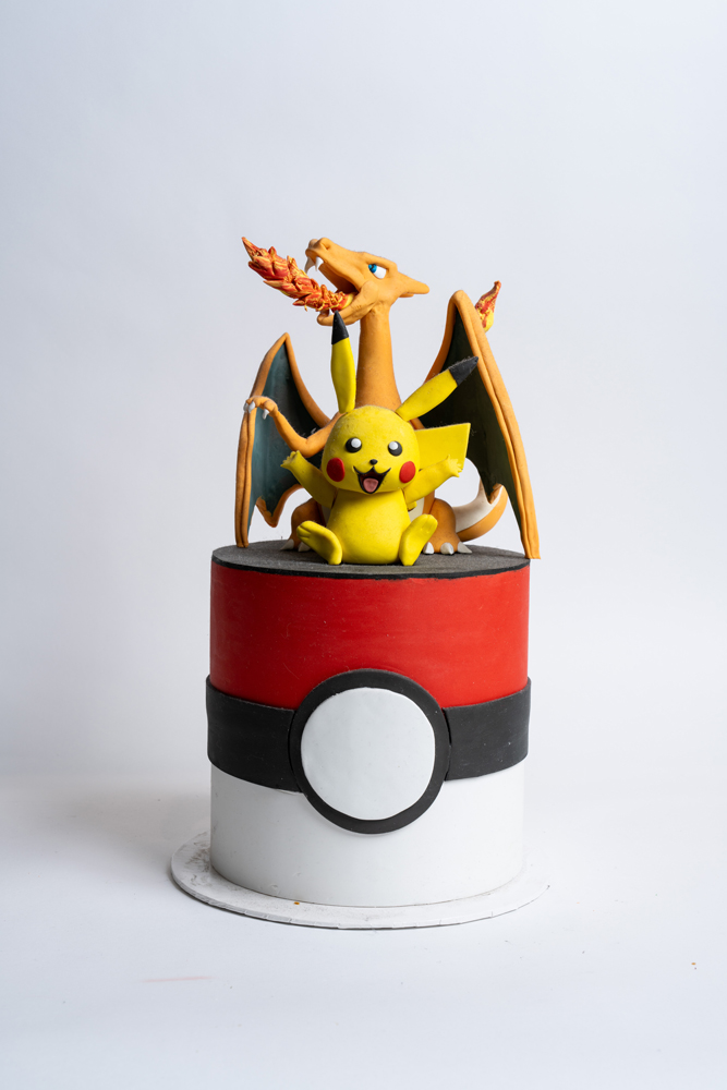 Custom Pokémon birthday cake from Mia's Bakery designed as a Pokéball with red, white, and black fondant layers. The cake is topped with detailed fondant figures of Pikachu sitting in front and Charizard with spread wings behind, holding its flame-tipped tail. The Pokéball features a white button center detail. Displayed against a white background.