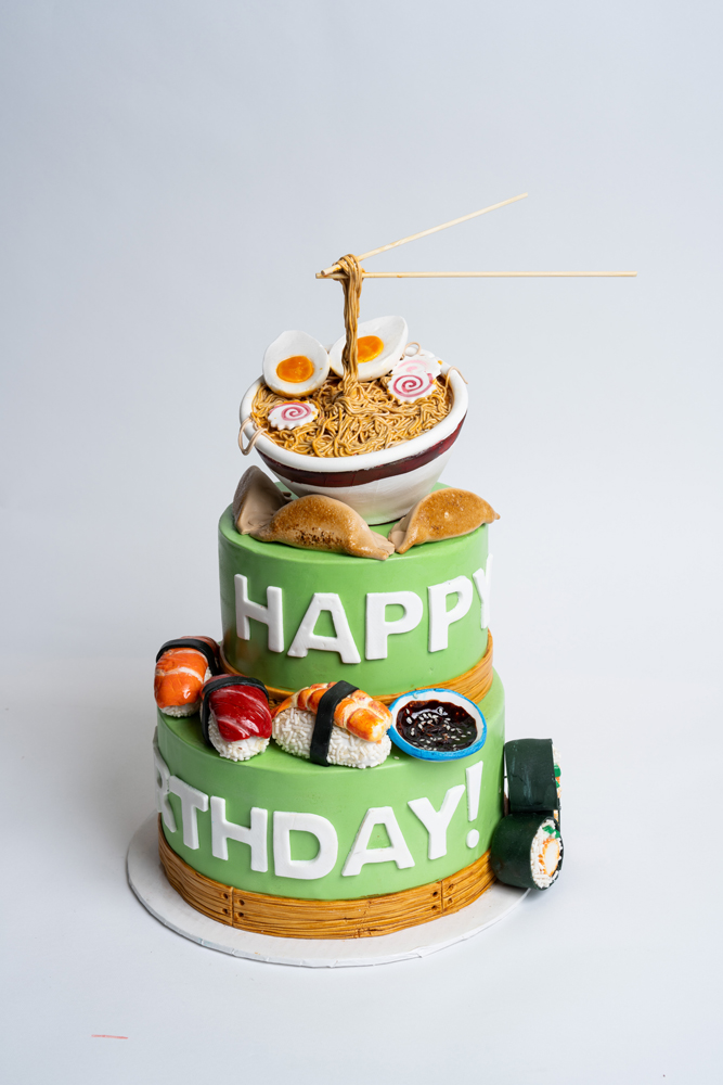 Custom Asian food-themed birthday cake from Mia's Bakery featuring a two-tier green cake with 'HAPPY BIRTHDAY!' text in white. The top displays a realistic ramen bowl with noodles, eggs, and naruto slices with chopsticks. The middle layer has edible gyoza dumplings, while the bottom tier is decorated with colorful fondant sushi rolls including salmon and tuna nigiri. Bamboo mat texture details the bottom edge. Displayed against a white background.