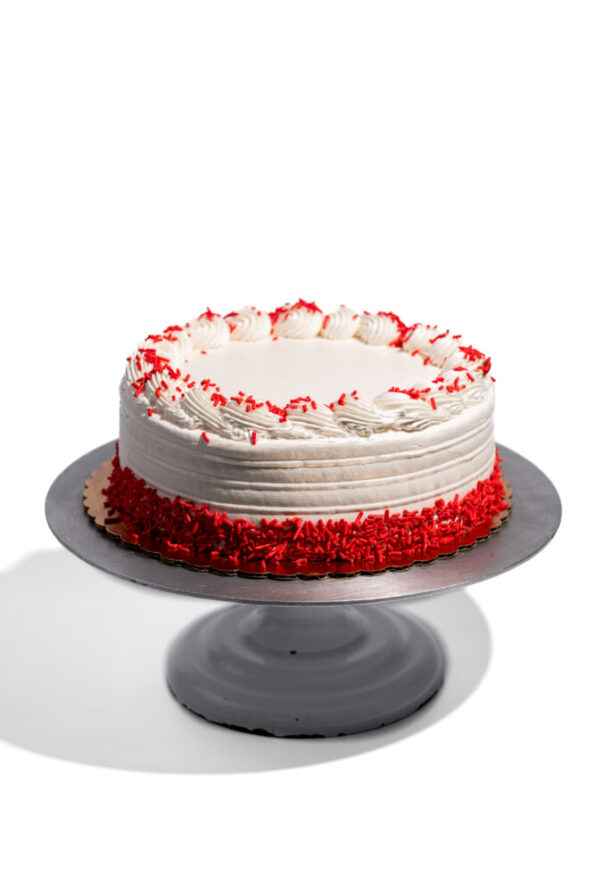 Custom vegan red velvet birthday cake from Mia's Bakery featuring a round single-layer cake with white vegan buttercream frosting and horizontal ridged texture on the sides. The cake is decorated with piped white rosettes around the top edge, each accented with red sprinkles. Red velvet cake crumbs coat the bottom edge. Displayed on a gray pedestal cake stand against a white background.