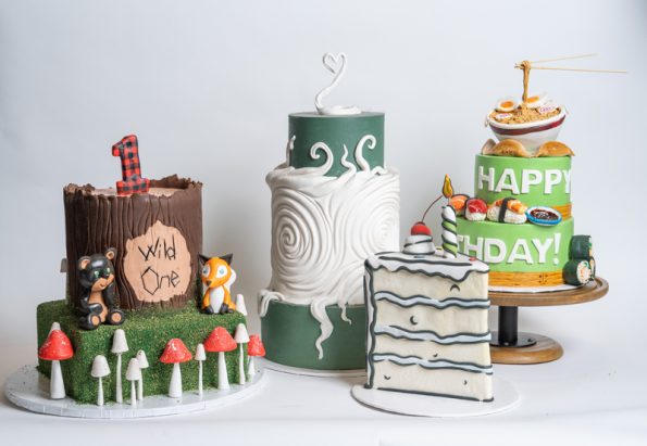 Custom themed birthday cake collection from Mia's Bakery featuring four unique designs: A woodland lumberjack cake with chocolate bark texture, mushrooms, and forest animals marked 'Wild One'; an elegant three-tier cake with green and white layers, wave patterns, and swan toppers; a nautical striped cake with wave design; and a colorful food-themed two-tier cake with 'HAPPY BIRTHDAY' text and miniature food decorations. All displayed on various stands against a white background.