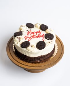 Oreo Birthday Cake from Mia's Bakery featuring a round chocolate layer cake with white vanilla buttercream on top and visible dark chocolate cake sides. The birthday cake is decorated with whole Oreo cookies arranged in a circle around the top edge, interspersed with piped white buttercream swirls and Oreo cookie crumb sprinkles. 'Happy Birthday!' is written in red cursive frosting in the center. Presented on a wooden pedestal cake stand against a white background.