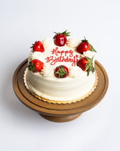Strawberry Shortcake Birthday Cake from Mia's Bakery featuring a round single-layer cake covered in smooth white vanilla buttercream with horizontal ridged texture on the sides. The birthday cake is decorated with alternating fresh strawberries and piped white buttercream rosettes around the top edge, with 'Happy Birthday!' written in red cursive frosting in the center. Presented on a wooden pedestal cake stand against a white background.