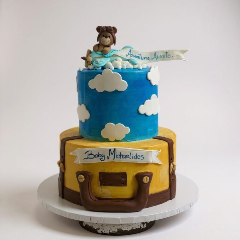 wo-tier adventure-themed baby shower cake. Bottom tier designed like a yellow suitcase with brown fondant straps and handle, top tier bright blue with white fondant clouds, topped with a fondant teddy bear flying a blue airplane and a banner