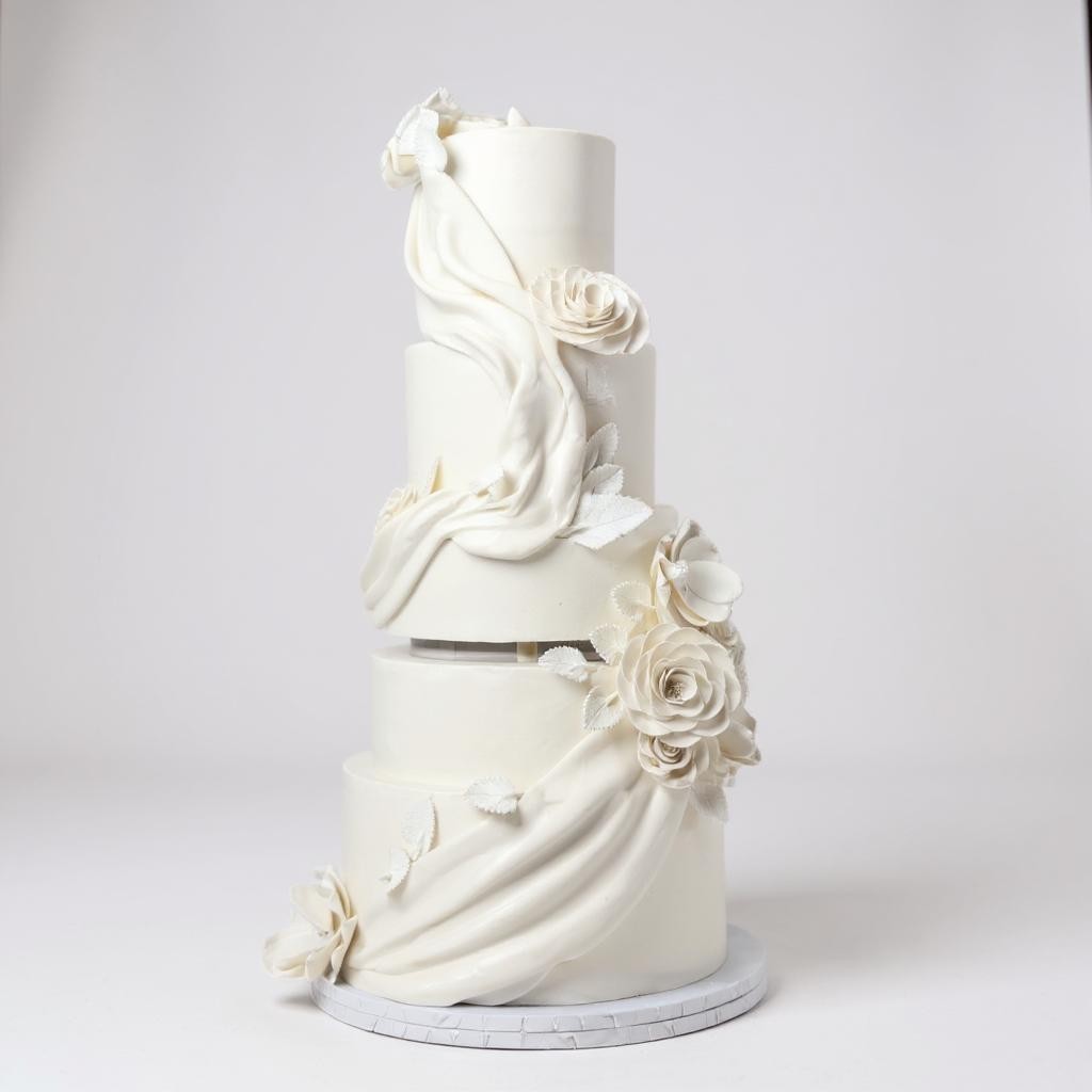 Four-tier white wedding cake adorned with cascading fondant drapes and elegant sugar roses, set on a white round cake stand against a plain light background